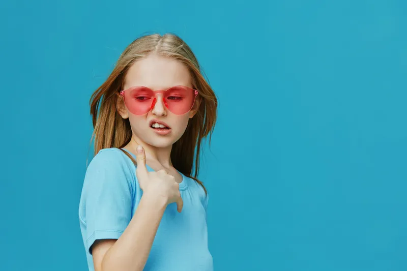 Young girl in trendy pink sunglasses with long blonde hair against a vibrant blue background, showing a playful and cool expression, ideal for fashion related content