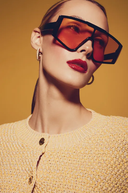 Stylish woman wearing oversized red sunglasses and yellow knitted sweater poses against warm yellow background. Fashion portrait with bold makeup and modern accessories.