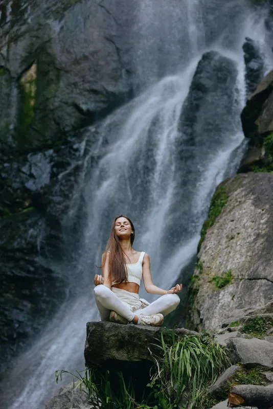 young woman meditating in lotus pose near waterfall wearing white activewear surrounded by rocks and greenery in nature forest setting