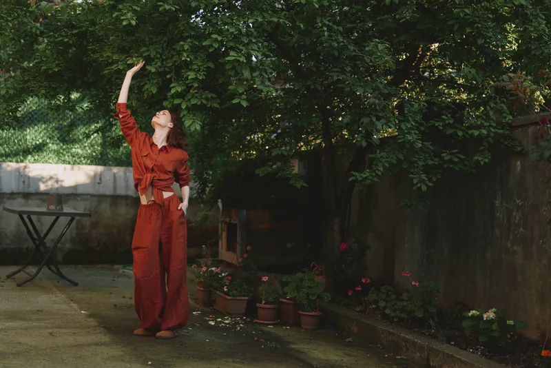 A stylish woman in a red jumpsuit reaches toward the sky in a sunlit garden, capturing a moment of playful confidence and outdoor lifestyle