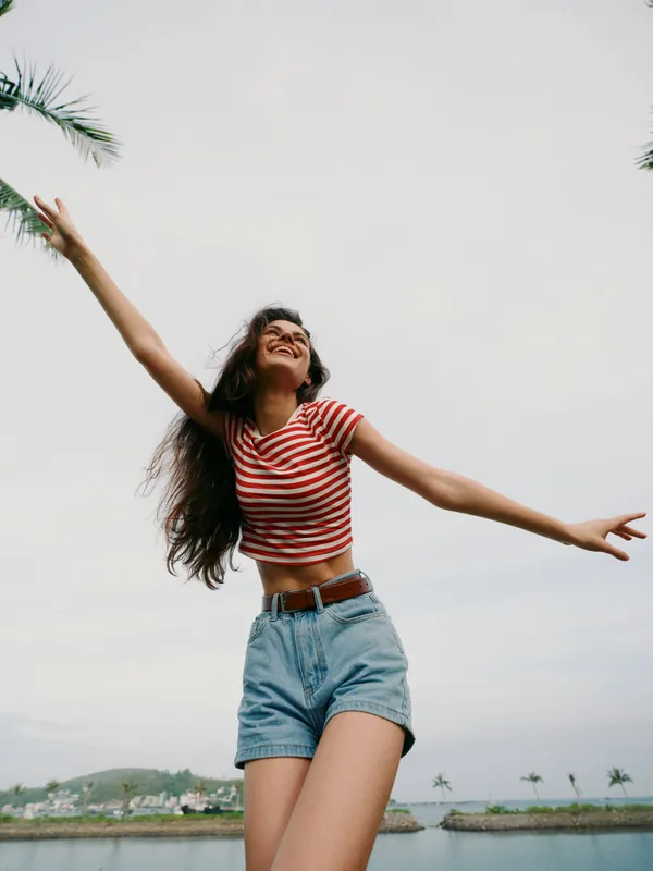 Joyful young woman stands outdoors with arms wide, wearing a red and white striped cropped top and denim shorts, mid movement under a bright open sky.
