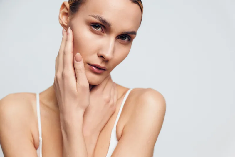 Close up of young woman with clear skin touching face gently looking at camera with calm expression isolated on white background natural beauty concept skincare and wellness.