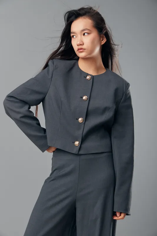 Young woman wearing a stylish gray suit with metallic buttons posing confidently with hand on hip against neutral gray background. Fashionable, modern, casual, and elegant portrait.
