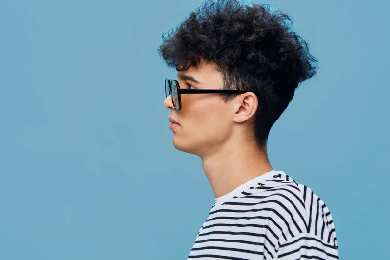 Young man with curly hair wearing glasses and a striped shirt stands in profile against a blue background, showcasing casual style and thoughtful expression.