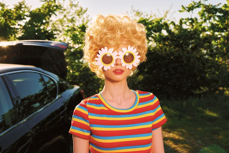 Young woman outdoors wears sunflower sunglasses and a colorful striped shirt, posing playfully with a bright, sunlit mood.
