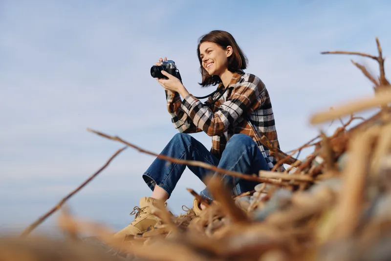 Photographer woman outdoors on a sandy beach, seated on driftwood, holding a camera and capturing landscape moments with a relaxed, focused expression in natural light.