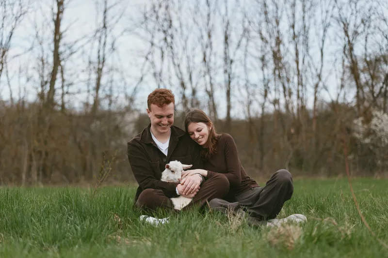 A couple sits in a grassy meadow with a goat kid, sharing a tender moment outdoors, capturing a lifestyle scene that highlights affection, family bonds, and rural simplicity.