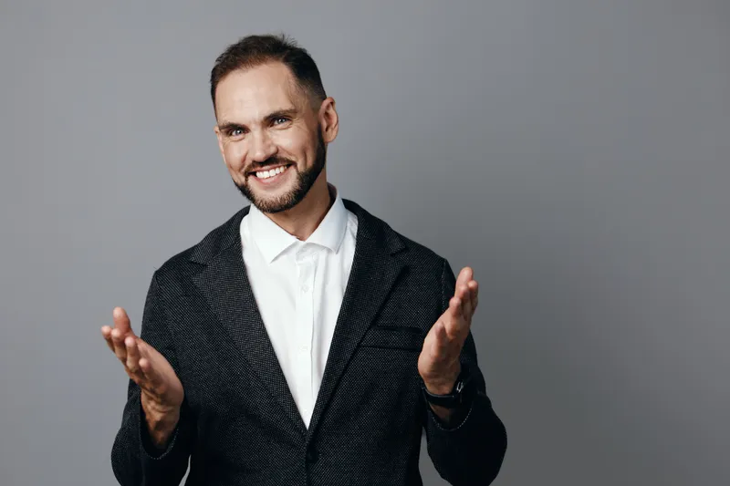 Confident businessman stands against a solid colored background, showing professional posture and calm emotion. He gestures with open hands, conveying friendly leadership and approachability in a