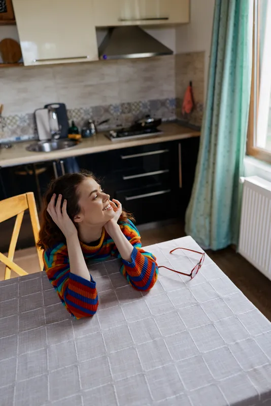Woman sits at kitchen table in a colorful striped sweater, hands to her head with a gentle smile, cozy home atmosphere, natural daylight from the window, relaxed vibe