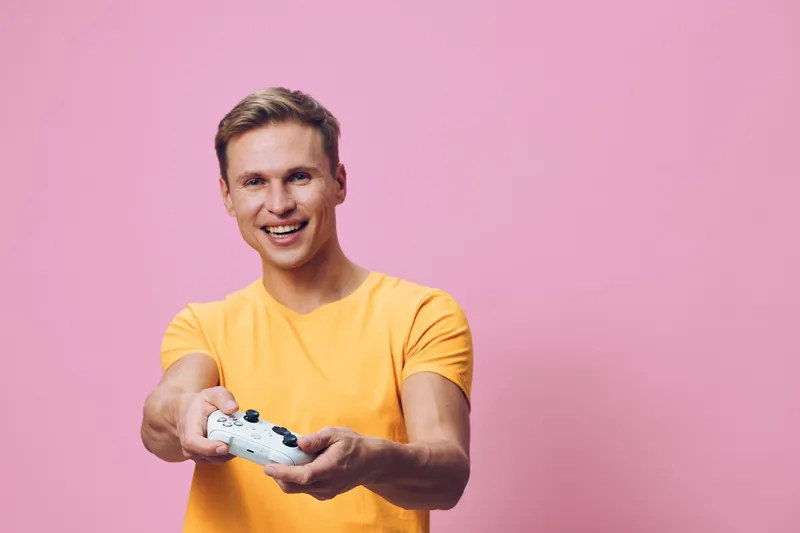 Smiling man wearing bright yellow t-shirt holding television remote control, looking at camera, cheerful expression, indoor studio shot with pink background, lifestyle concept.