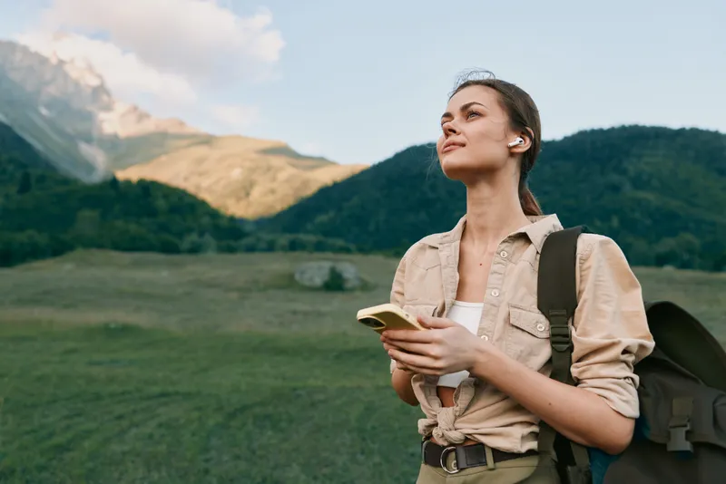 A young woman with a backpack holds a smartphone while standing in a green mountainous landscape under a partly cloudy sky. Concept for outdoor lifestyle and travel.