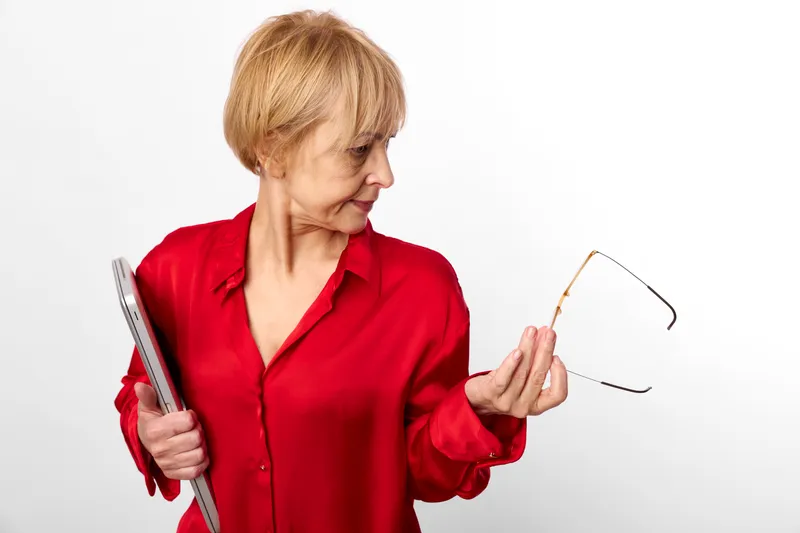 Older woman in red shirt holding book and glasses, studio white background, focused puzzled expression, looking at notes, soft light, modern everyday life calm