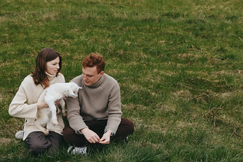 A calm outdoor scene features a couple with a goat kid on lush grass, sharing a quiet moment of affection and bonding, celebrating rural lifestyle, companionship, and the tender connection between