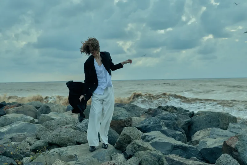 Individual on rugged coastal rocks, wind swept, wearing a white shirt and light pants, adjusting a dark jacket as waves crash and dramatic clouds roll above