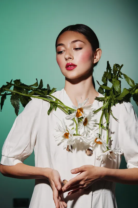 Young woman in white dress holds chamomile flowers and green leaves on shoulders. Creative beauty and fashion portrait with natural elements on light green background.