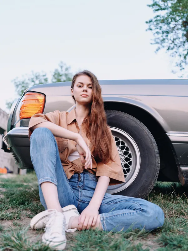 Young woman in casual outfit sitting near vintage car, expressing confidence and relaxation in a natural outdoor setting, embodying summer vibes and freedom.