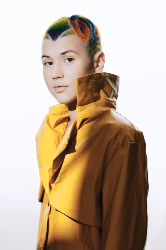 Woman with colorful short hair wearing a yellow trench coat, posing against a clean white background with a serious and modern style.