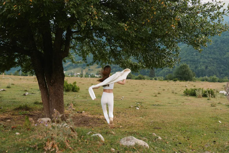 Young woman dancing in a white outfit twirls beside a large tree in an open field under a bright sky, conveying a sense of freedom and playful movement.