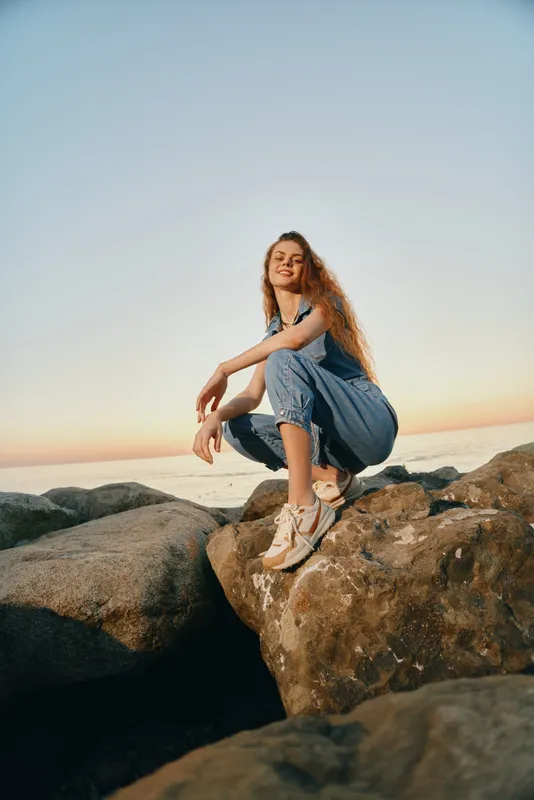 lifestyle woman in modern boho-western denim street style crouching on rocks with warm film color tones during sunset outdoors