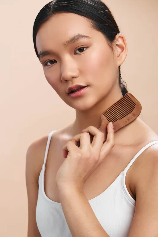 Asian woman portrait with a wooden comb near her neck, soft lighting, minimal makeup, and a casual white top, presenting a calm, intimate grooming moment.