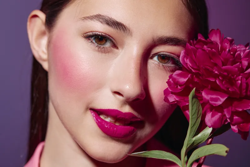 Young woman with vibrant pink lipstick poses closely holding a deep magenta flower against a purple background. Creative beauty and fashion portrait with trendy makeup.
