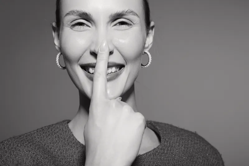 Smiling white woman with pale skin holds finger vertically near nose in black and white portrait on plain background concept for silence and positivity.
