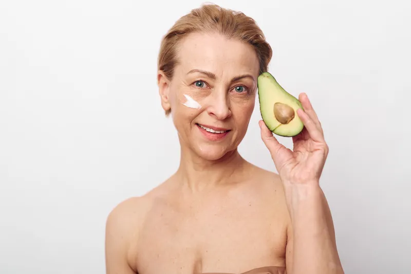 Mature woman with natural skin holds fresh avocado near face, applying cream on cheek. Concept of skincare, healthy lifestyle, natural beauty, and organic facial care for aging skin.