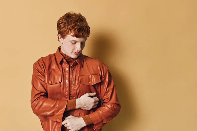 Young man with red hair buttoning up a brown leather jacket standing against beige background. Casual fashion, portrait and style in autumn clothing for youth and confidence.