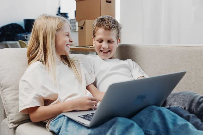 Two children sitting on a sofa with a laptop, smiling and enjoying time together in a cozy living room. Concept of family bonding, technology use, and leisure activities at home.
