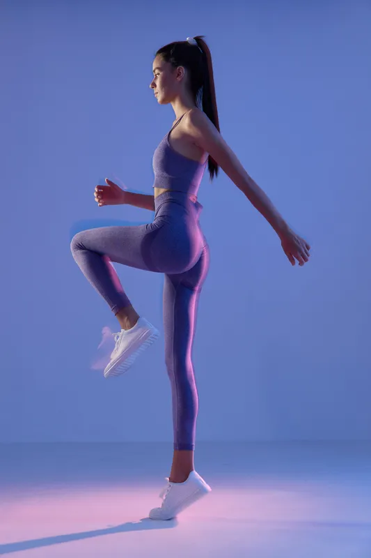Athletic woman sport fitness performing high knee exercise in purple activewear and white sneakers against purple gradient background, showing strength and focus.