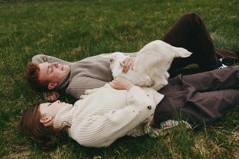Young couple lies on grass outdoors enjoying time together with a playful baby goat in a relaxed lifestyle scene during autumn.