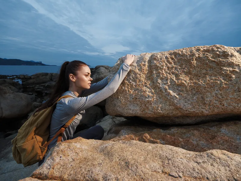 Young female climber wearing gray sportswear with a backpack overcoming a rock formation by the sea under a cloudy sky, showcasing determination and adventure
