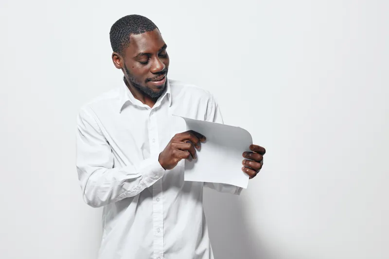 African American man with dark skin wearing white shirt holding and looking at blank paper sheet on white background for presentation or communication concept in studio.