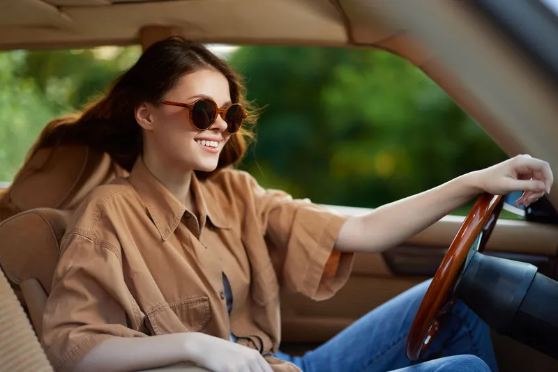 Young woman in stylish sunglasses and oversized shirt enjoying a summer drive in a vintage car amidst green foliage.