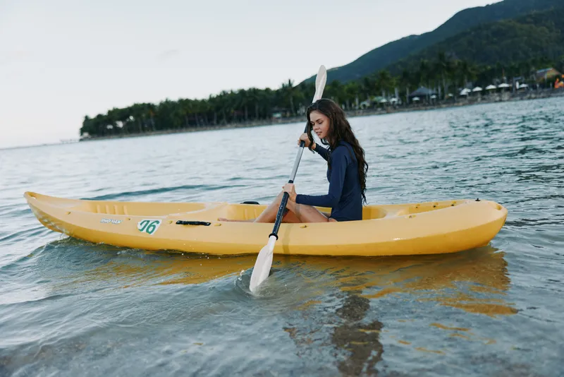 A lone kayaker paddles a yellow sit-on-top kayak on calm water, near shore with green hills in the distance, presenting a serene outdoor adventure moment.