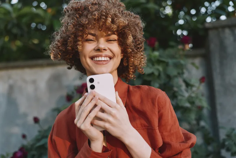 Cheerful woman outdoors, smiling with smartphone in hand, casual rust shirt, sunlit moment, natural colors, leafy background, relaxed lifestyle, candid portrait, warm breeze, joyful mood