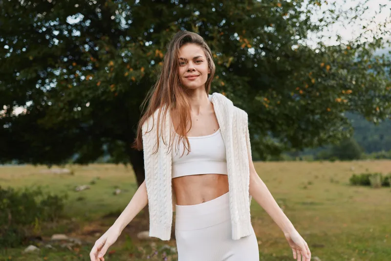 Smiling woman in a white crop top and knit vest in a sunlit field. Natural outdoor setting, relaxed lifestyle, fresh air, warm and welcoming mood and carefree calm.