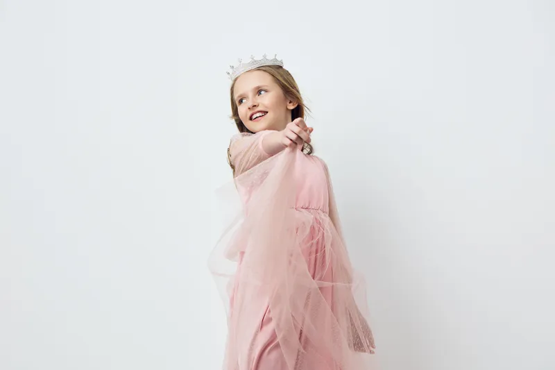 Cute girl in a pink princess dress with a crown, smiling and pointing, set against a light background, perfect for children s parties and celebrations