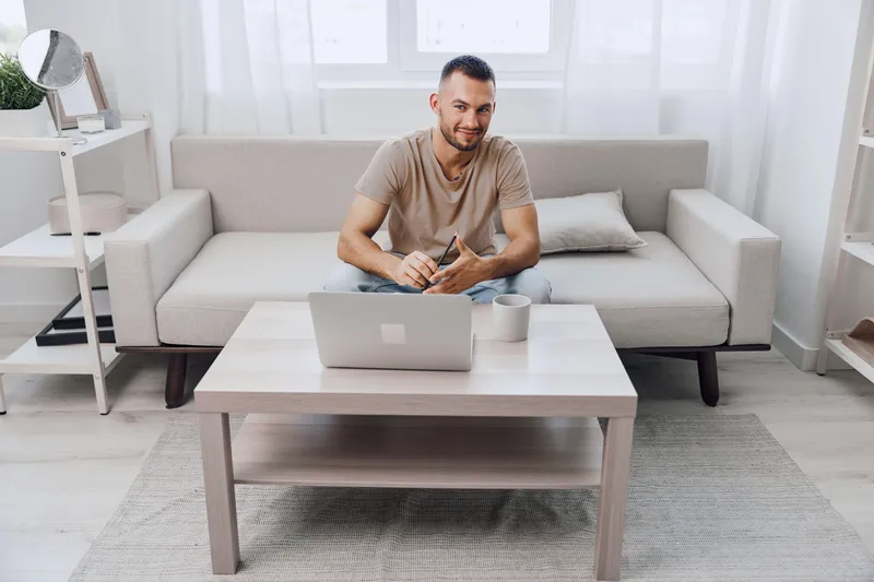 Young man working on a laptop in a modern living room, showcasing a cozy atmosphere with natural light and minimalistic decor, inspiring productivity and comfort.