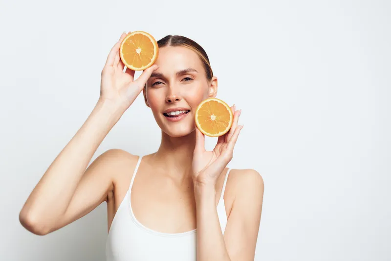 A smiling young woman in a white tank top holds two orange halves near her face against a white background, representing fresh fruit, healthy lifestyle, and natural beauty concepts.