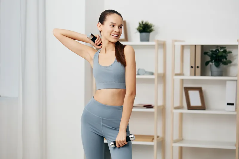 Young woman doing fitness workout at home, wearing matching activewear and holding dumbbells while stretching her shoulder, smiling confidently in a bright minimalist living room with shelves.