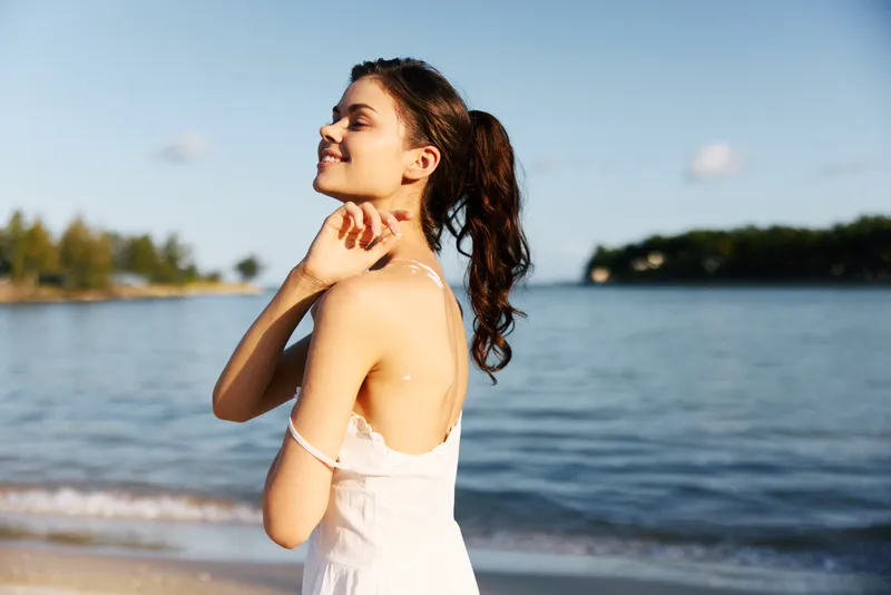 Portrait of a young woman standing on a sunlit beach, wearing a white dress, hair tied back, a gentle smile, and relaxed pose as soft waves kiss the shore.