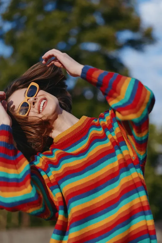 Young woman smiling in sunglasses and a striped sweater outdoors, joyful mid twenties model adjusting retro glasses in sunlit green park, casual outfit and colorful fashion portrait.