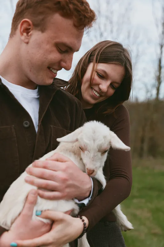 Joyful couple outdoors cuddle a young goat kid, sharing affection and smiles in a rural setting with grass and trees, capturing a warm, natural lifestyle moment of companionship.