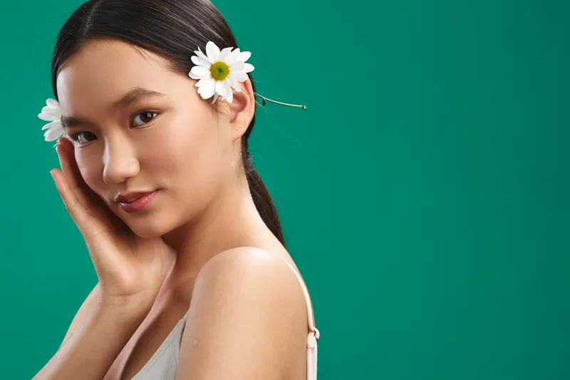 asian woman with white daisies in her hair poses against a vivid green backdrop, showcasing calm beauty, soft lighting, and a fresh natural vibe that highlights gentle features and a serene mood.