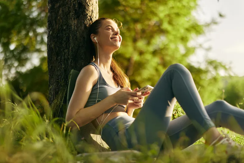 Young woman smiling while listening to music in nature, wearing sportswear, relaxed and happy in a sunny outdoor setting, surrounded by greenery