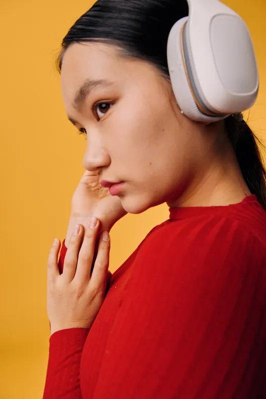 Young woman wearing wireless headphones listens thoughtfully with hands near face, dressed in red long sleeve top against yellow background. Technology and music concept with side profile view.