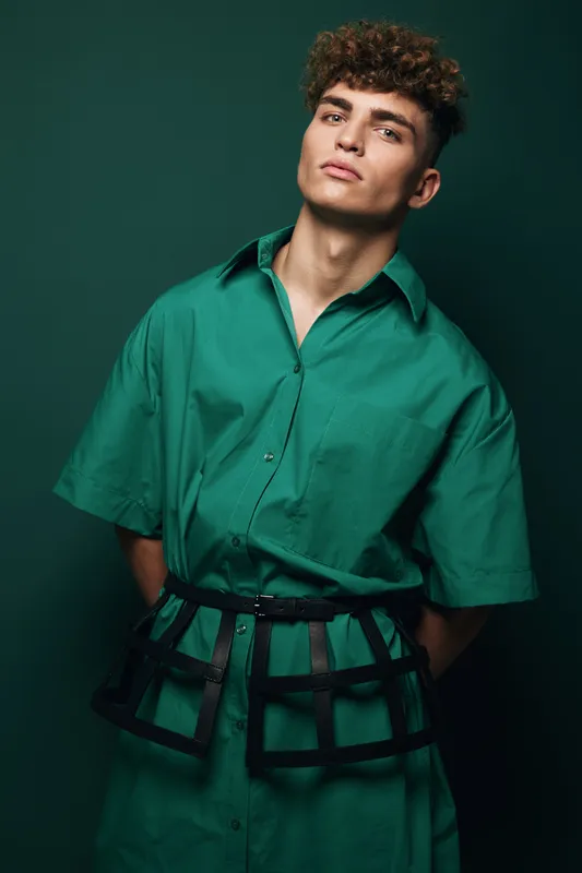 Young man with curly hair wearing oversized green shirt and black fashion accessory posing in studio, trendy style and unique outfit for modern casual fashion shoot with confident expression.