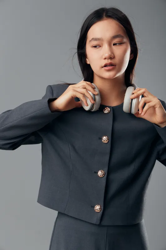 Young woman in gray tailored suit holding gray headphones against gray background. Portrait of professional female with straight hair in stylish outfit for technology and fashion concept.