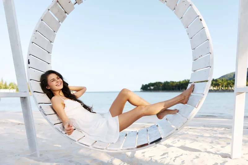 A sunny beach scene shows a relaxed woman in a white dress lounging on a circular wooden swing by the calm sea, enjoying warm light and gentle ocean breeze.